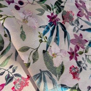 May include: A close-up of a white fabric with a floral print. The fabric features pink, purple, and green flowers, as well as blue leaves. The fabric is textured and has a subtle sheen.