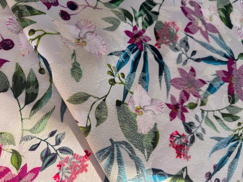 May include: A close-up of a white fabric with a floral print. The fabric features pink, purple, and green flowers, as well as blue leaves. The fabric is textured and has a subtle sheen.