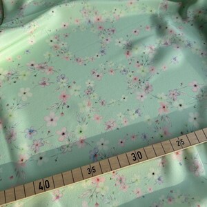 May include: A close-up of a light green fabric with a delicate floral pattern. The fabric is measured with a wooden ruler, showing measurements in inches.
