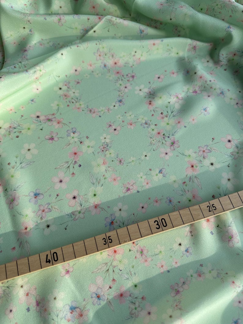 May include: A close-up of a light green fabric with a delicate floral pattern. The fabric is measured with a wooden ruler, showing measurements in inches.