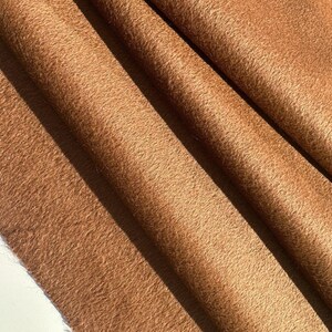 May include: Close-up of a folded, textured fabric in a warm, earthy brown hue. The material has a soft, fuzzy appearance, with visible folds and shadows that add depth. The fabric's texture suggests a plush or woolen material.