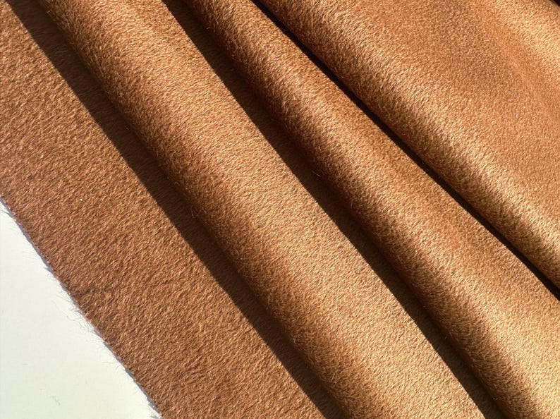 May include: Close-up of a folded, textured fabric in a warm, earthy brown hue. The material has a soft, fuzzy appearance, with visible folds and shadows that add depth. The fabric's texture suggests a plush or woolen material.