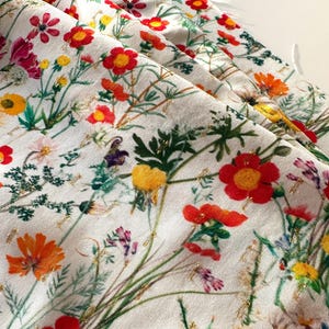May include: White fabric with a floral print featuring red, yellow, and green flowers with gold accents.