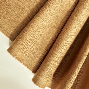 May include: A piece of tan fabric with a distinct, raised, wavy, and textured surface pattern. The material is folded into several vertical pleats, creating deep shadows and highlights across the fabric's surface, showcasing its unique tactile quality.