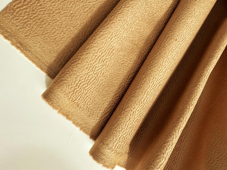 May include: A piece of tan fabric with a distinct, raised, wavy, and textured surface pattern. The material is folded into several vertical pleats, creating deep shadows and highlights across the fabric's surface, showcasing its unique tactile quality.