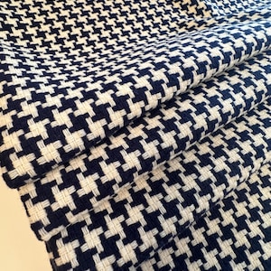 May include: Close-up of a folded fabric with a classic houndstooth pattern. The fabric features a repeating design of navy blue and white shapes, creating a textured appearance. The material is tightly woven, suggesting durability and a tailored look.