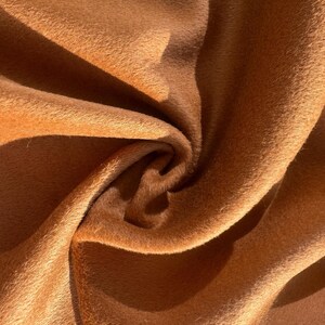 May include: Close-up of a luxurious, textured fabric in a warm, golden-brown hue. The material is soft and plush, with visible folds and shadows that create depth and dimension. The fabric's surface has a slight sheen.