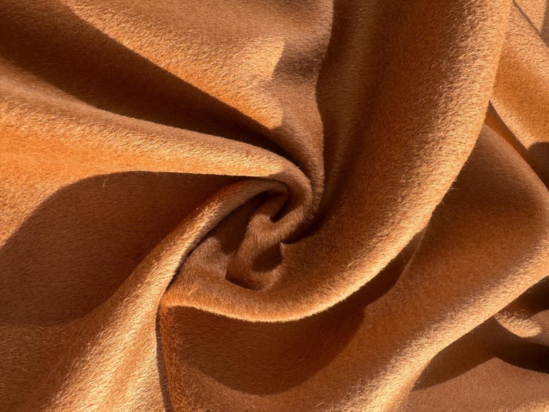 May include: Close-up of a luxurious, textured fabric in a warm, golden-brown hue. The material is soft and plush, with visible folds and shadows that create depth and dimension. The fabric's surface has a slight sheen.