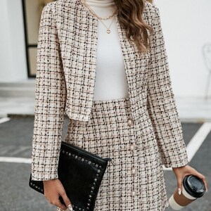 Puede incluir: Un traje de falda de tweed con una chaqueta corta y una falda con botones de perla. La falda y la chaqueta son de un estampado de cuadros beige y marr&oacute;n.