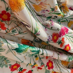May include: White fabric with a floral print featuring red, yellow, and pink flowers with green leaves and gold accents.