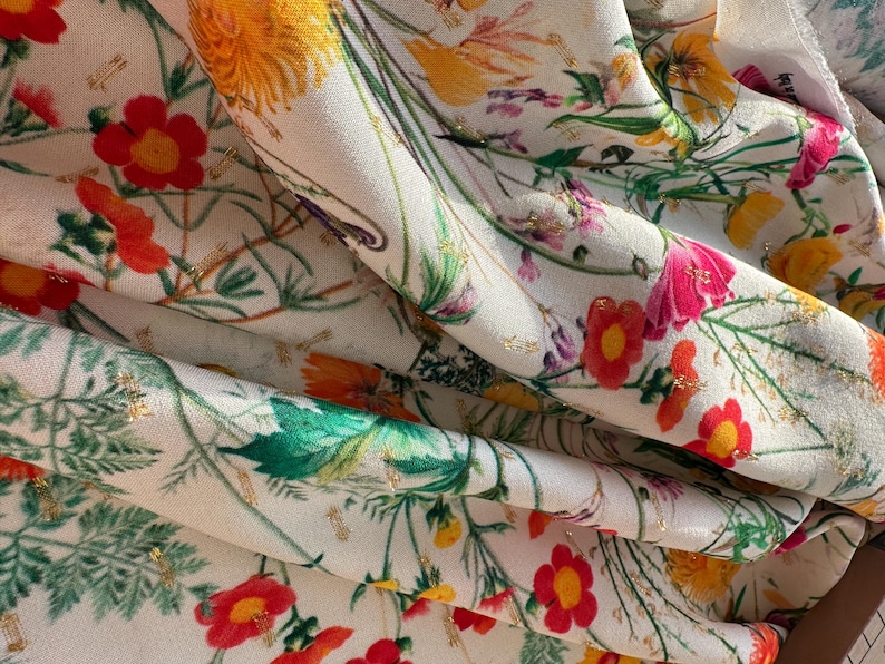 May include: White fabric with a floral print featuring red, yellow, and pink flowers with green leaves and gold accents.