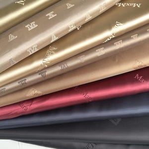 Puede incluir: Una pila de tela en varios colores, incluyendo dorado, beige, burdeos, azul marino y negro. La tela tiene un brillo sutil y está impresa con el logotipo "MaxMara". La tela es probablemente para ropa o accesorios de moda.