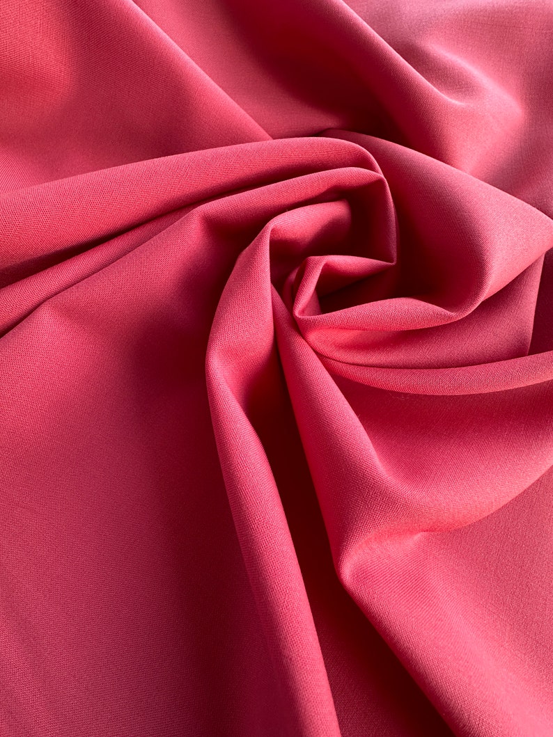 Op de afbeelding: Een close-up van een gladde, zachte, roze stof met een subtiele textuur. De stof is gevouwen en gedrapeerd, waardoor een vloeiende, elegante uitstraling ontstaat.