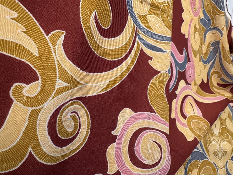May include: A close-up of a fabric with a burgundy background and a swirling pattern of gold, pink, and blue.