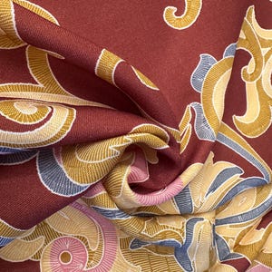 May include: Red fabric with a swirling pattern in shades of gold, cream, and blue. The fabric has a textured surface and a luxurious feel.
