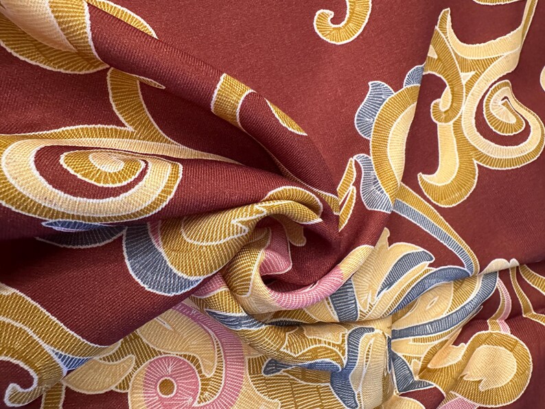 May include: Red fabric with a swirling pattern in shades of gold, cream, and blue. The fabric has a textured surface and a luxurious feel.