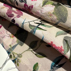 May include: Close-up of a beige fabric with a floral pattern. The fabric features pink, purple, green, and blue flowers and leaves. The fabric has a textured surface.