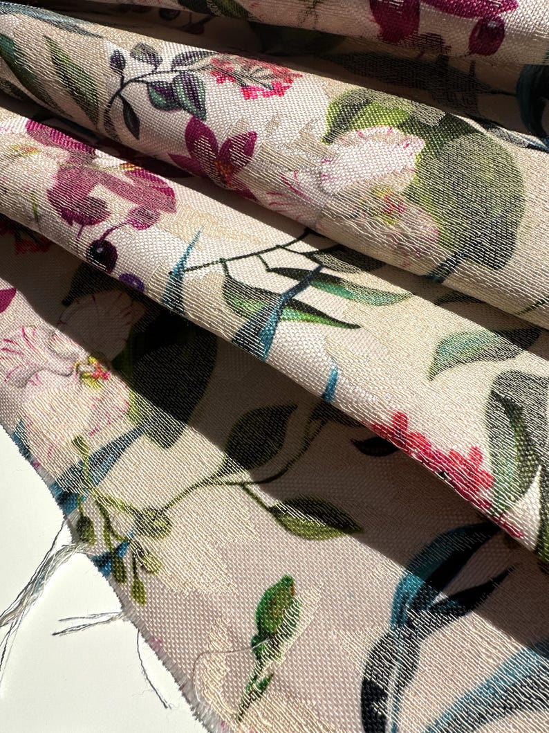 May include: Close-up of a beige fabric with a floral pattern. The fabric features pink, purple, green, and blue flowers and leaves. The fabric has a textured surface.