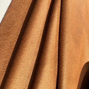May include: A piece of tan-colored fabric with a soft, suede-like texture. The material is draped in vertical folds, creating deep shadows and highlights across the surface, emphasizing the matte finish and the consistent, warm brown color.
