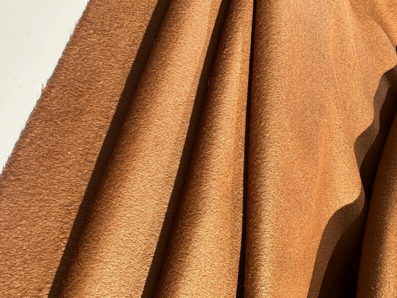 May include: A piece of tan-colored fabric with a soft, suede-like texture. The material is draped in vertical folds, creating deep shadows and highlights across the surface, emphasizing the matte finish and the consistent, warm brown color.