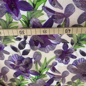 May include: Purple and green floral fabric with a white background. The fabric has a textured surface and a repeating pattern of flowers and leaves.