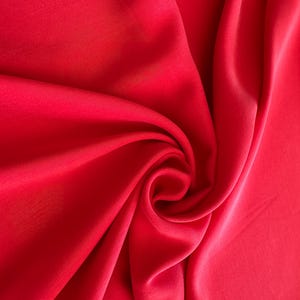 May include: A close-up of a smooth, red fabric draped in soft folds. The fabric has a subtle sheen and a luxurious feel.