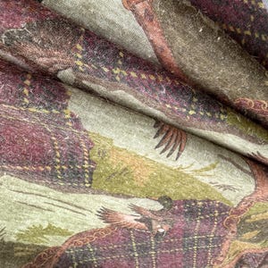 May include: Close-up of a fabric with a detailed print featuring a hunting scene. The design incorporates elements of plaid in shades of burgundy, brown, and green. The fabric appears to be soft and textured, with visible folds and creases.