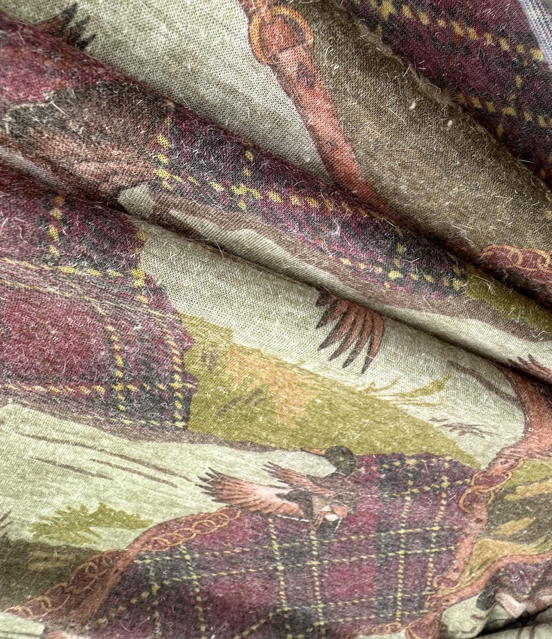May include: Close-up of a fabric with a detailed print featuring a hunting scene. The design incorporates elements of plaid in shades of burgundy, brown, and green. The fabric appears to be soft and textured, with visible folds and creases.