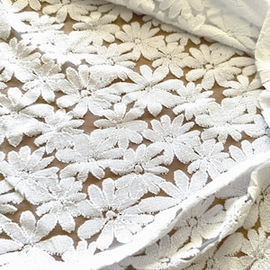 Könnte beinhalten: Weißer Spitzenstoff mit Gänseblümchenmuster. Der Stoff ist gefaltet und zeigt das komplizierte florale Design. Die Spitze ist mit erhabenen, strukturierten Blütenblättern und Blättern versehen, was ein zartes und luftiges Aussehen erzeugt. Der Hintergrund ist eine hellbraune Oberfläche.