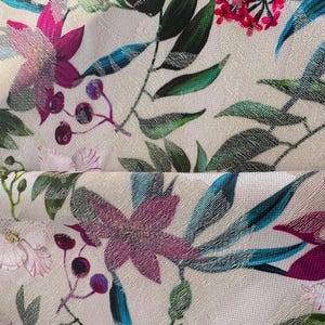 May include: A white fabric with a floral print featuring pink, purple, and blue flowers. The fabric has a textured surface and a subtle sheen.