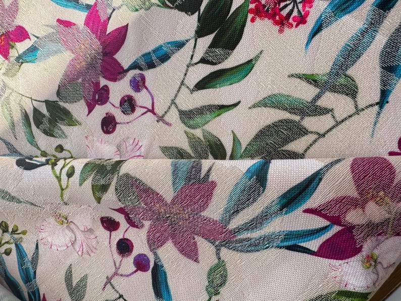 May include: A white fabric with a floral print featuring pink, purple, and blue flowers. The fabric has a textured surface and a subtle sheen.