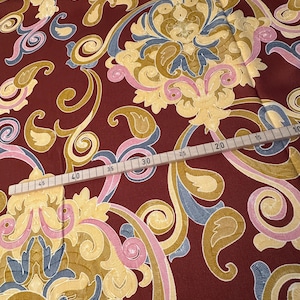 May include: Red fabric with a gold, pink, and blue paisley pattern. A wooden ruler with markings in inches is visible at the bottom of the image.