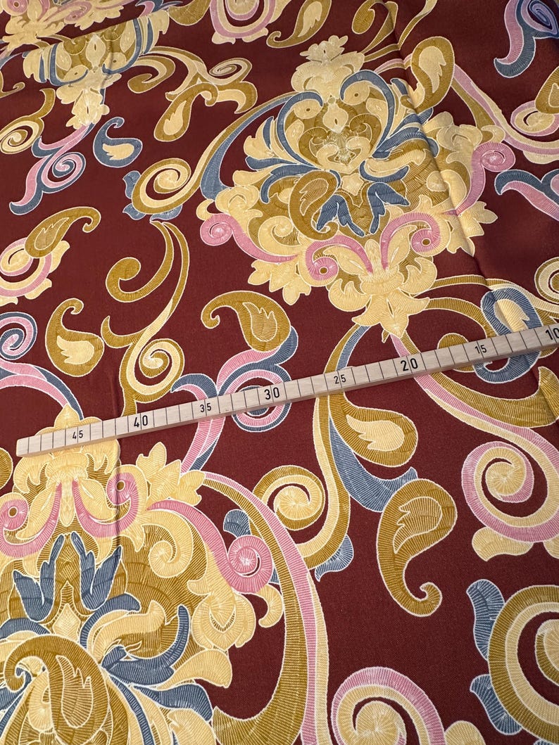 May include: Red fabric with a gold, pink, and blue paisley pattern. A wooden ruler with markings in inches is visible at the bottom of the image.