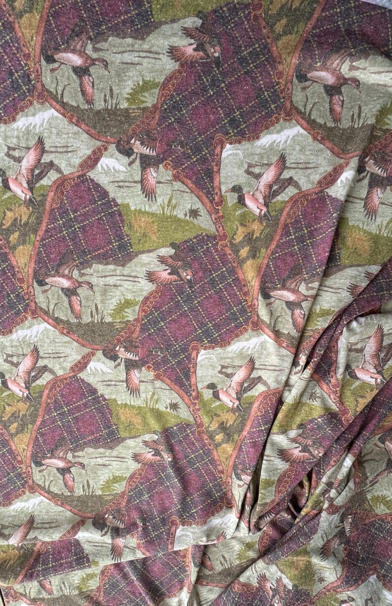 May include: Fabric with a repeating pattern of flying ducks, plaid-like shapes, and a landscape background in shades of green, brown, and burgundy. The fabric has a vintage or rustic aesthetic.