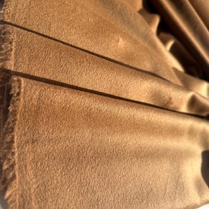 May include: A length of solid brown fabric with a soft, brushed texture and a matte finish. The material features a distinct nap and visible, slightly frayed edges along the side, showing a dense, uniform color throughout the draped folds.