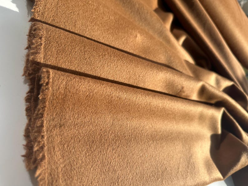 May include: A length of solid brown fabric with a soft, brushed texture and a matte finish. The material features a distinct nap and visible, slightly frayed edges along the side, showing a dense, uniform color throughout the draped folds.