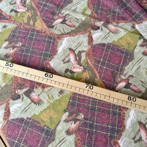 May include: Fabric with a repeating pattern of flying ducks, plaid sections in burgundy and brown, and a light green background. The design includes a gold chain-like border. A wooden ruler is placed across the fabric, measuring from 50 to 80 inches.