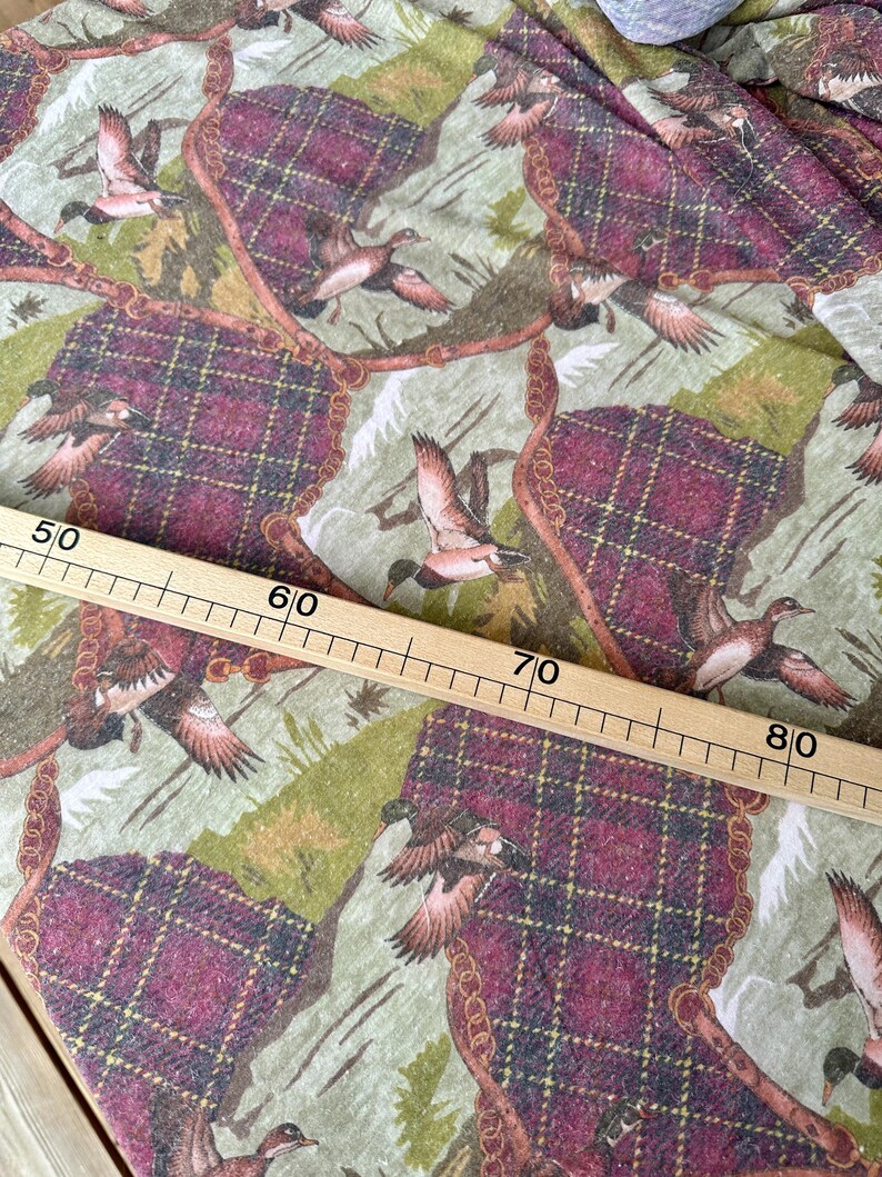 May include: Fabric with a repeating pattern of flying ducks, plaid sections in burgundy and brown, and a light green background. The design includes a gold chain-like border. A wooden ruler is placed across the fabric, measuring from 50 to 80 inches.