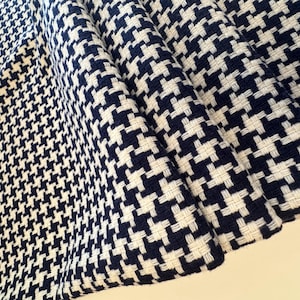 May include: Close-up of a folded fabric with a classic houndstooth pattern. The fabric features a repeating design of navy blue and white woven checks. The texture of the fabric is visible, with the weave creating a slightly raised surface.