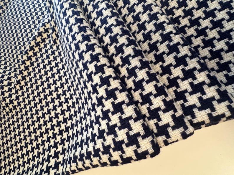 May include: Close-up of a folded fabric with a classic houndstooth pattern. The fabric features a repeating design of navy blue and white woven checks. The texture of the fabric is visible, with the weave creating a slightly raised surface.