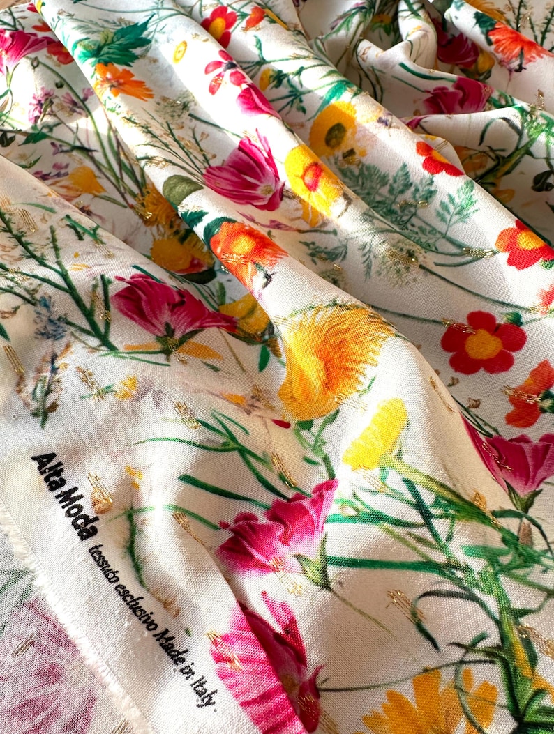 May include: White fabric with a floral print featuring pink, yellow, and red flowers. The fabric has a gold thread woven into the design. The fabric has a label that reads "Alta Moda tessuto esclusivo Made in Italy."