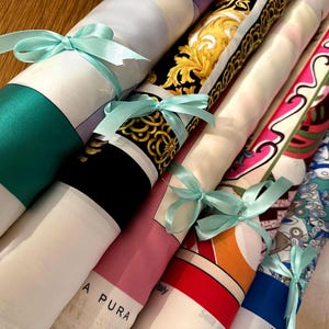 Op de afbeelding: Gerolde zijden sjaals vastgebonden met lichtblauwe linten. De sjaals hebben verschillende patronen en kleuren, waaronder goud, zwart, roze, rood en blauw. Sommige sjaals hebben de tekst "in Italy" en "A Pura".
