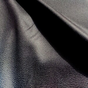 May include: A dark charcoal gray fabric with a distinctively pebbled, granular texture. The material is draped to create deep, shadowed folds that highlight the uneven surface and the matte finish of the textile, showing light and dark contrast.
