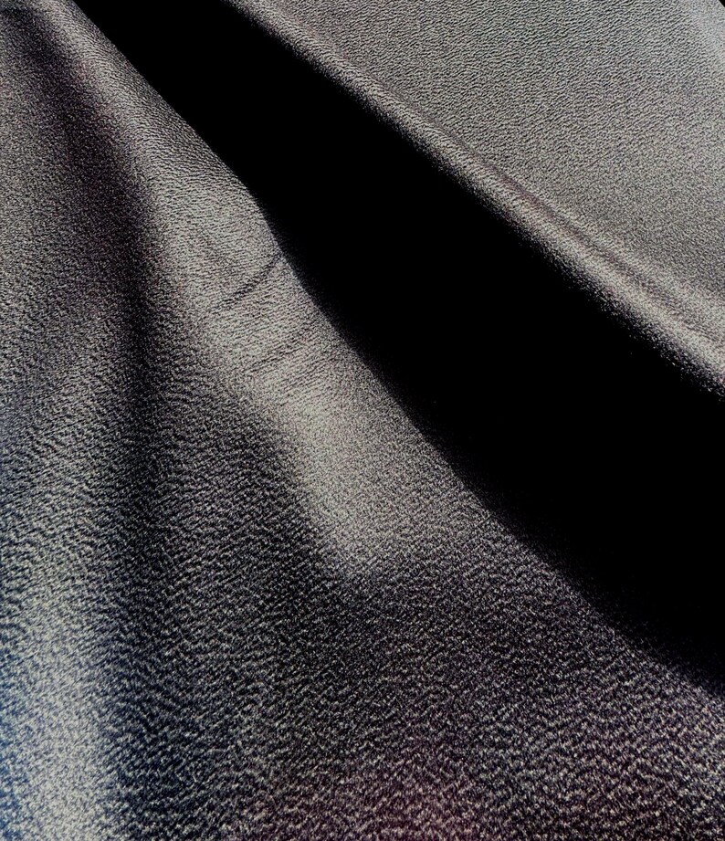 May include: A dark charcoal gray fabric with a distinctively pebbled, granular texture. The material is draped to create deep, shadowed folds that highlight the uneven surface and the matte finish of the textile, showing light and dark contrast.