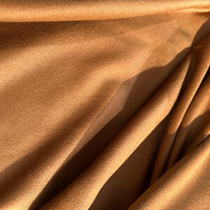 May include: Close-up of a soft, textured fabric in a warm, golden-brown hue. The material is draped, creating folds and shadows that add depth and dimension. The fabric's surface appears plush and inviting.