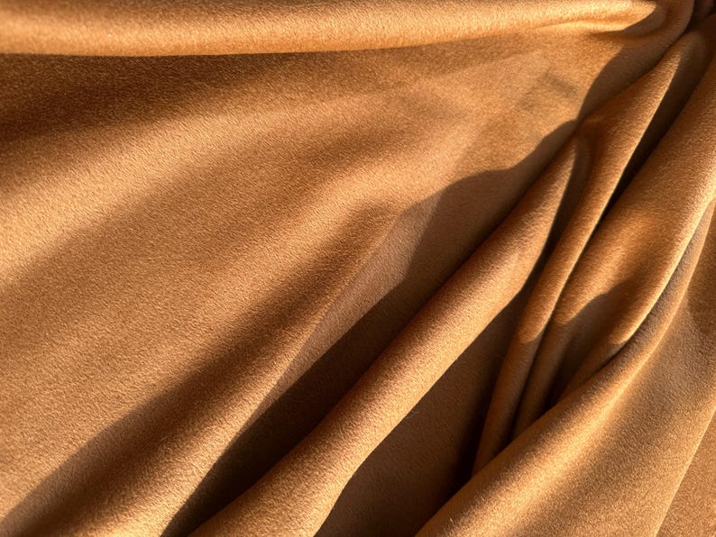 May include: Close-up of a soft, textured fabric in a warm, golden-brown hue. The material is draped, creating folds and shadows that add depth and dimension. The fabric's surface appears plush and inviting.