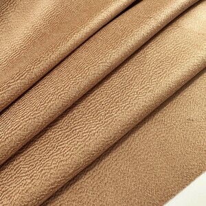 May include: A piece of fabric in a solid light brown color featuring a distinct, fine, and wavy textured surface. The material is folded into several diagonal layers, creating deep shadows and highlights that emphasize the rippled pattern.