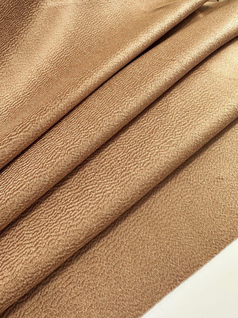 May include: A piece of fabric in a solid light brown color featuring a distinct, fine, and wavy textured surface. The material is folded into several diagonal layers, creating deep shadows and highlights that emphasize the rippled pattern.
