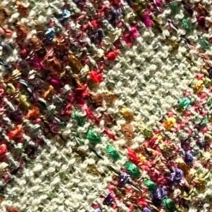 Puede incluir: Primer plano de una tela tejida con una superficie texturizada. La tela es de color beige claro con hilos multicolores tejidos en toda su longitud. Los hilos son en tonos de rosa, rojo, verde, azul y dorado.