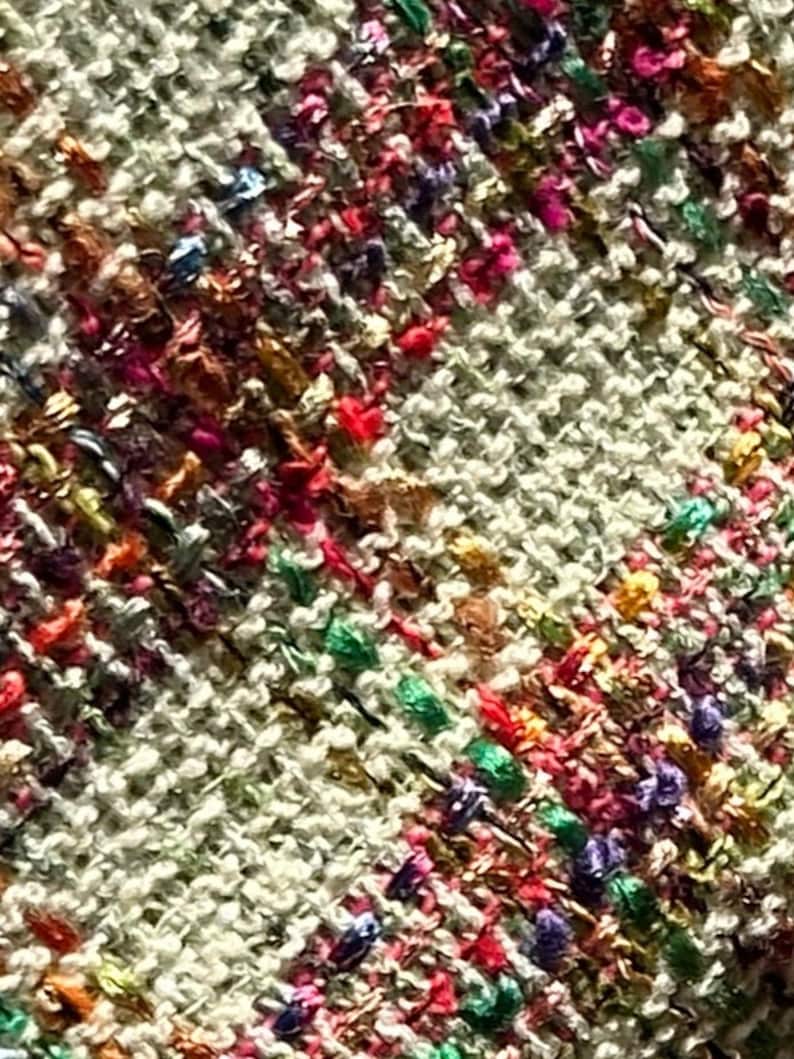 Puede incluir: Primer plano de una tela tejida con una superficie texturizada. La tela es de color beige claro con hilos multicolores tejidos en toda su longitud. Los hilos son en tonos de rosa, rojo, verde, azul y dorado.